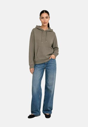 Donna che indossa una felpa verde opaco con cappuccio e tasca frontale, jeans blu a gamba larga e scarpe nere basse, in piedi con una mano in tasca.