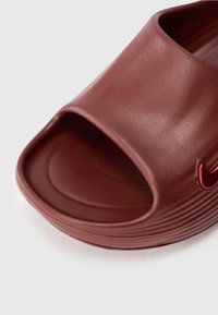 Κοντινό πλάνο από μια μπορντό παντόφλα με υφασμάτινη σόλα και ένα μικρό, γυαλιστερό κόκκινο λογότυπο στο πλάι.