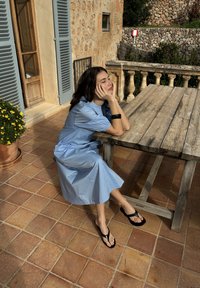Jeune femme en robe bleue et sandales noires assise sur une terrasse carrelée, reposant sa tête sur sa main à côté d'une table en bois.