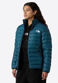 Giovane donna che guarda a destra, indossando una giacca trapuntata di colore verde scuro sopra un felpa nera, entrambe con il logo "The North Face", in piedi davanti a uno sfondo bianco.