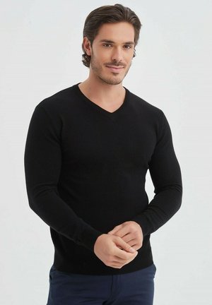 Pull noir ajusté à col V avec des manches longues. Texture lisse et poignets côtelés, conçu pour un ajustement près du corps. Le modèle porte un pantalon marine.