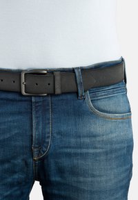 Une ceinture noire avec une finition texturée, dotée d'une boucle rectangulaire en argent. Portée avec un jean en denim bleu et une chemise blanche.