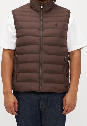 Homme portant une veste matelassée marron zippée par-dessus une chemise blanche à manches courtes et un jean bleu foncé, debout devant un fond uni.