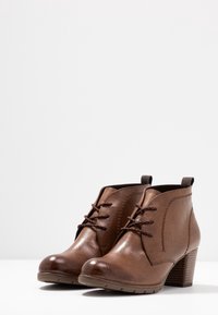 Brązowe skórzane botki za kostkę o teksturowanym wykończeniu, zawiązywane z przodu, z obcasem o wysokości 7 cm. Posiadają język do zakupu i gumową podeszwę zapewniającą przyczepność.