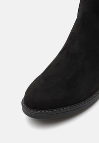 Botte en daim noir vue de face, avec une pointe arrondie et des coutures visibles le long du bord de la semelle en caoutchouc sur fond blanc.