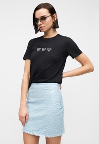 Γυναίκα που φοράει μαύρο μπλουζάκι με τρεις καρδιές από στρας και ανοιχτό μπλε μίνι φούστα με υφή, στέκεται με το ένα χέρι στη μέση, φοράει γυαλιά ηλίου.