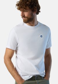 T-shirt bianco in cotone con scollo rotondo e maniche corte, caratterizzato da un piccolo logo blu nell'area sinistra del petto. Texture liscia.