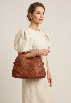 Woman wearing a cream puff-sleeve dress holding a large brown leather handbag with floral embossing and gold studs.