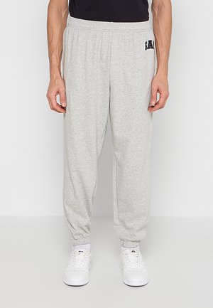Homme portant un pantalon de survêtement gris clair avec des poignets élastiques et des baskets blanches, debout devant un fond clair uni.
