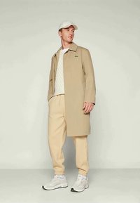 Νεαρός άνδρας φορώντας μπεζ παλτό, κρεμ πουλόβερ με πλέξη, μπεζ παντελόνι, λευκά αθλητικά παπούτσια και λευκό καπέλο, που στέκεται μπροστά σε απλό φόντο.