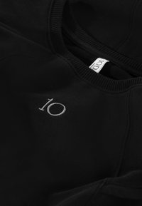 Schwarzer Sweatshirt mit strukturiertem Stoff, versehen mit einer aufgestickten "10" in silbernem Faden auf der Vorderseite und einem gewebten Etikett am Hals.