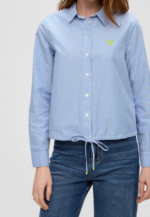 Vrouw draagt een blauw-wit gestreept overhemd met knopen en kraag, gestrikt in de taille, gecombineerd met een blauwe spijkerbroek.