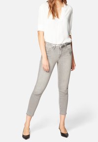 Vaqueros skinny grises de tiro medio, con un diseño de tachuelas en el bolsillo derecho, combinados con una blusa blanca y tacones negros.
