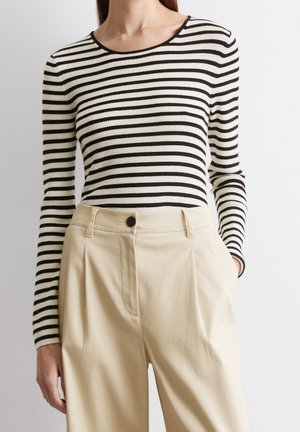 Kvinde iført sort-hvid stribet bluse med lange ærmer, stoppet ned i højtaljede beige bukser med plisseringer og knaplukning.