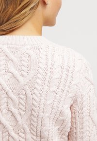 Pull rose clair en maille avec des motifs torsadés complexes, un col rond et des fibres texturées. Vue arrière mettant en valeur les coutures détaillées.
