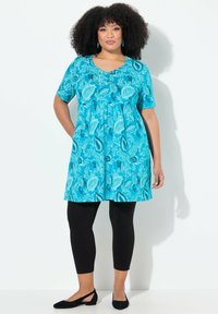 Turkusowa sukienka w kwiatowy wzór, z krótkimi rękawami i podkreśloną talią, w połączeniu z czarnymi legginsami i czarnymi płaskimi butami.