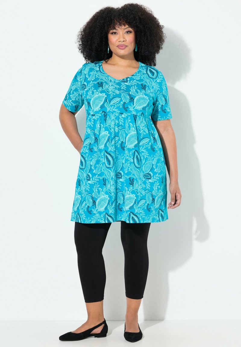 Turkusowa sukienka w kwiatowy wzór, z krótkimi rękawami i podkreśloną talią, w połączeniu z czarnymi legginsami i czarnymi płaskimi butami.