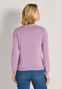 Frau mit kurzem blonden Haar, die einen helllila gestrickten Pullover und blaue Jeans trägt, steht mit dem Rücken zu einem schlichten Hintergrund.