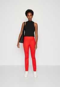 Donna in piedi con un top senza maniche nero, pantaloni rossi brillanti, sneakers bianche e una borsa a spalla nera contro uno sfondo chiaro e semplice.