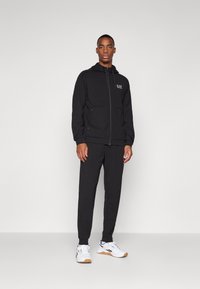 Black zip-up hoodie with two front pockets and logo, paired with matching black pants. Model wears white athletic shoes with gum sole.