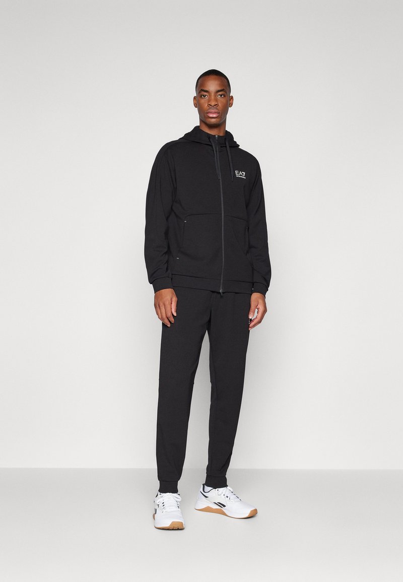 Black zip-up hoodie with two front pockets and logo, paired with matching black pants. Model wears white athletic shoes with gum sole.