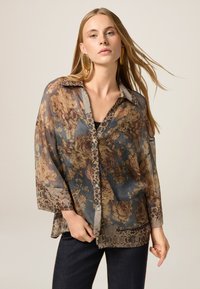 Blusa floreale trasparente con tonalità beige e marrone, dotata di bottone frontale, colletto e maniche con motivo. Abbinata a jeans scuri.