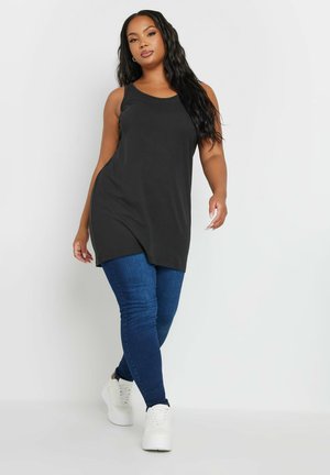 Donna con capelli lunghi e scuri che indossa una tunica nera senza maniche, jeans blu e scarpe da ginnastica bianche, che cammina su uno sfondo bianco.