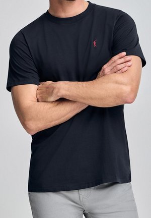 Mężczyzna w czarnej koszulce z krótkim rękawem z małym czerwonym logo gracza polo, z założonymi rękami, ubrany w jasnoszare spodnie.