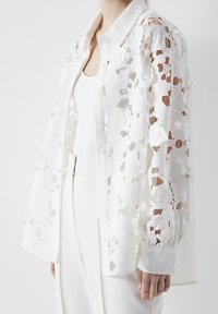 White lace shirt with floral patterns, sheer sleeves, and a button-up front. Fabric is light and textured, paired with a white tank top underneath.