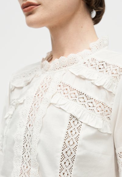 Γυναίκα που φοράει λευκό πουκάμισο με περίτεχνα δαντελένια πάνελ, βολάν περιγράμματος και ψηλό δαντελένιο γιακά με κυματοειδές σχέδιο.