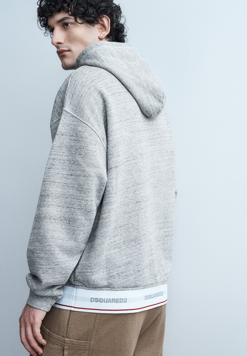 Giovane con capelli ricci che indossa una felpa grigia con cappuccio e pantaloni marroni, di profilo su uno sfondo grigio chiaro.