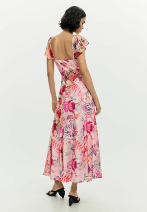 Kobieta o krótkich włosach, ubrana w różową suknię maxi w kwiaty z falbankowymi rękawami i odkrytymi plecami, stojąca na jednolitym tle.