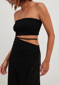 Černý bezramínkový top se střihovým designem a diagonálním lemem. Obsahuje boční výřezy a zlaté kovov�é akcenty. Hladká, elastická tkanina.