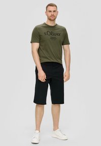 T-shirt vert olive avec texte noir, associé à un short noir à genoux. Le modèle porte des baskets blanches. Design simple et décontracté.