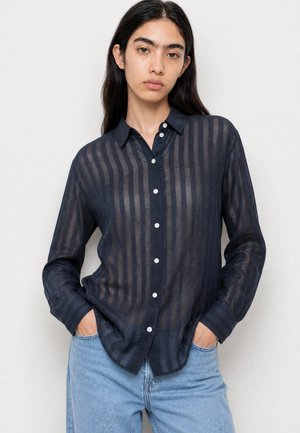 Jeune femme aux longs cheveux foncés portant une chemise boutonnée à rayures bleu marine transparente et un jean bleu clair, les mains dans les poches.