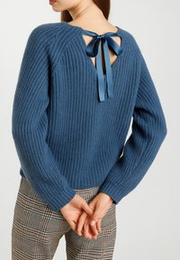 Maglione blu a coste con apertura sulla schiena legata da un nastro in satin. Tessuto texture e vestibilità comoda. Abbinato a pantaloni a quadri.