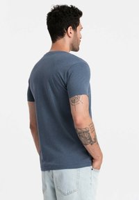 Tričko s krátkým rukávem z tmavě modré bavlny, s kulatým výstřihem a hladkou texturou. Na pravém rameni se nacházejí viditelné tetování.
