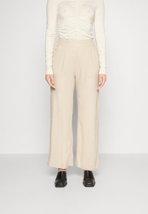 Frau trägt beige hoch taillierte weit geschnittene Hose, cremefarbenes langärmeliges gerafftes Oberteil und schwarze Schuhe mit quadratischer Spitze, steht vor einfachem Hintergrund.