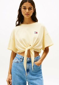 T-shirt giallo a maniche corte con nodo frontale, realizzata in morbido cotone, caratterizzata da una piccola etichetta con logo. Abbinata a jeans in denim blu a vita alta.