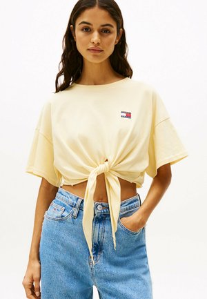 Korte mouwen, gele T-shirt met knoopdetail, gemaakt van zacht katoen, met een klein logo patch. Gepaard met hoge, blauwe spijkerbroek.
