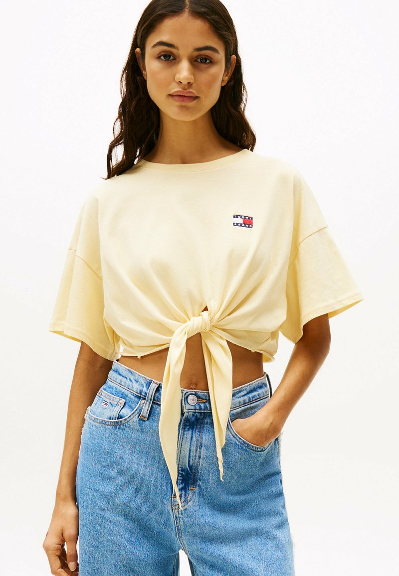 T-shirt giallo a maniche corte con nodo frontale, realizzata in morbido cotone, caratterizzata da una piccola etichetta con logo. Abbinata a jeans in denim blu a vita alta.