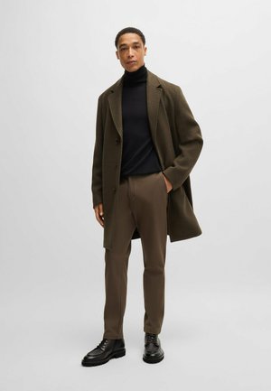 Olijfgroen wollen blendjas, zwarte coltrui, bruine op maat gemaakte broek en zwarte veterschoenen, staand tegen een lichtgrijze achtergrond.