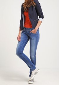 Blue denim skinny jeans with a mid-rise fit, faded detailing, and five-pocket design. Paired with a navy blazer and casual sneakers.