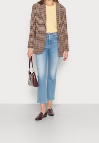 Blazer à carreaux marron et crème sur un pull jaune, jeans cropped bleus et mocassins bordeaux, tenant un sac gris texturé.
