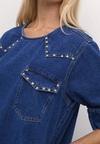 Blå denimtop med kort ärm, två fickor med rhinestonapplikationer och rundad halsringning med dekorativ sömnad.