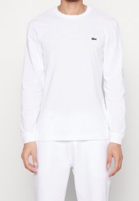 Vit långärmad skjorta i bomull med rund hals och en liten grön Lacoste-logotyp på vänster bröst. Slät textur, avslappnad passform.