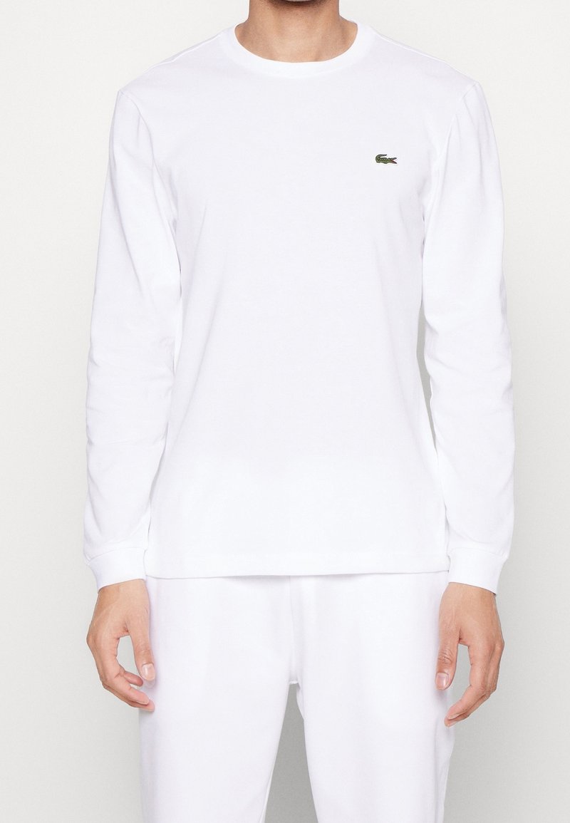 Vit långärmad skjorta i bomull med rund hals och en liten grön Lacoste-logotyp på vänster bröst. Slät textur, avslappnad passform.