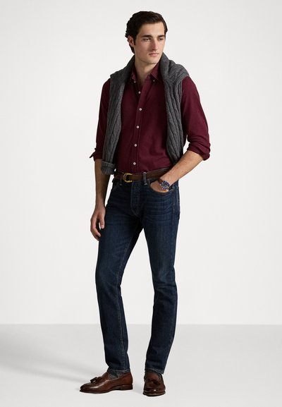 Chemise boutonnée bordeaux associée à un jean bleu foncé. Pull en maille torsadée gris drapé sur les épaules. Chaussures marron avec des détails.