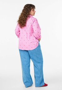 Rosa gestreiftes Hemd mit roten Herzmustern, lange Ärmel, lockere Passform, kombiniert mit hellblauen Jeans mit weitem Bein und roten Schuhen.