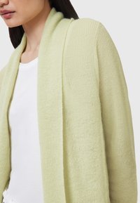 Cardigan verde chiaro, lavorato a maglia in modo largo, con una finitura testurizzata e design aperto, abbinato a una maglietta bianca attillata sotto.